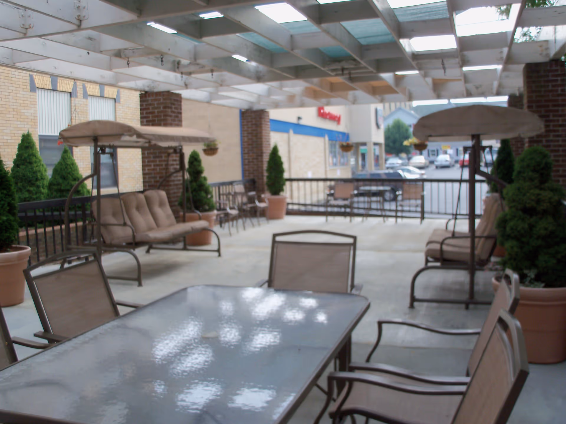 Covered outdoor patio with a glass-top table, metal chairs, cushioned swing seats and potted plants under a pergola overlooking a parking area.