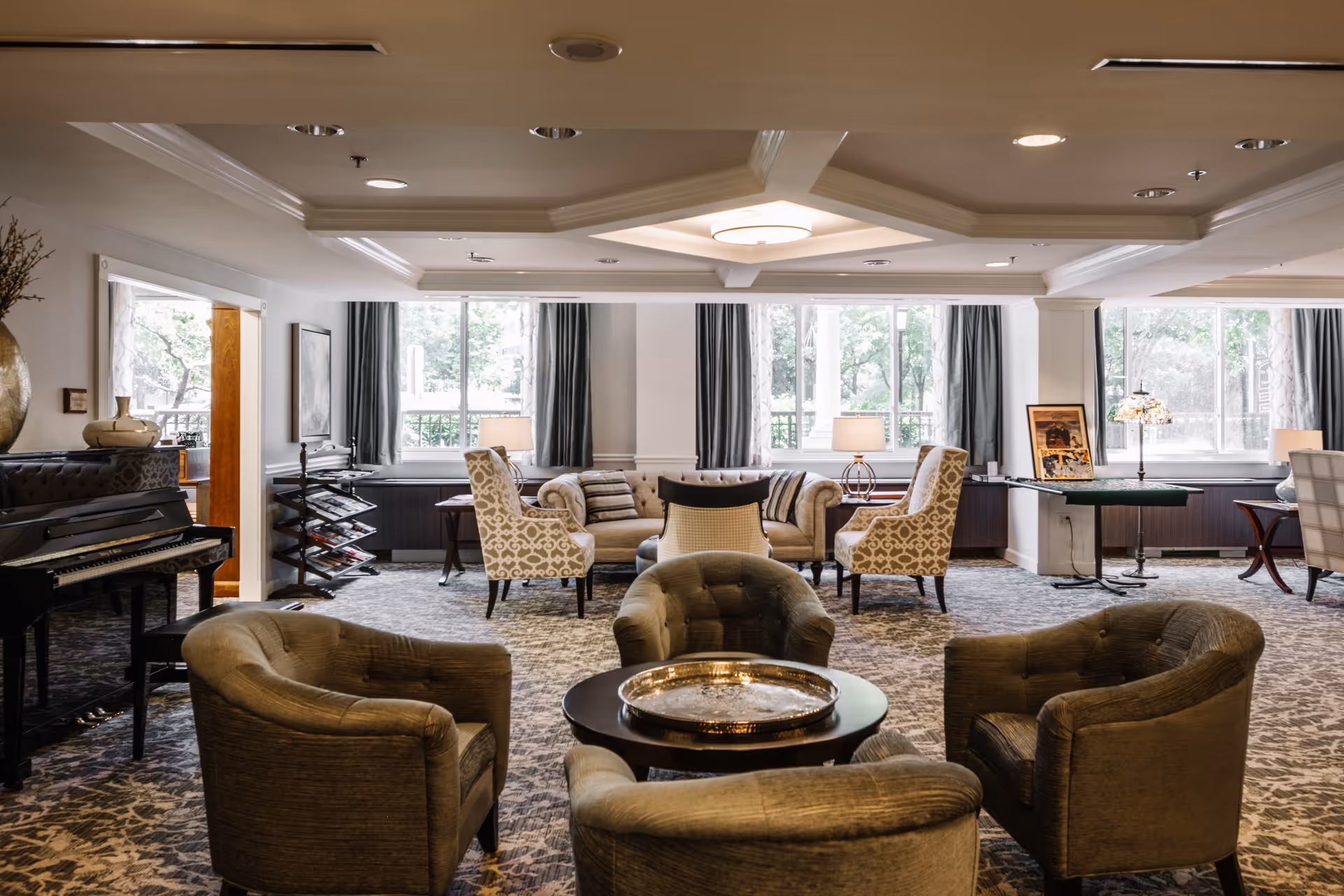 A spacious and well-lit living room area in Thomas House Retirement Community featuring a set of four cushioned armchairs arranged around a round coffee table with a decorative tray. In the background, there is a beige sofa flanked by two patterned armchairs, large windows with gray curtains letting in natural light, a piano on the left side, and a small table with a lamp and framed pictures on the right.