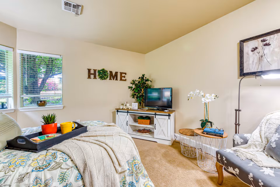 Bright furnished bedroom with a bed, tray with cups, TV on a white stand, armchair, plants, and 'HOME' wall decor.