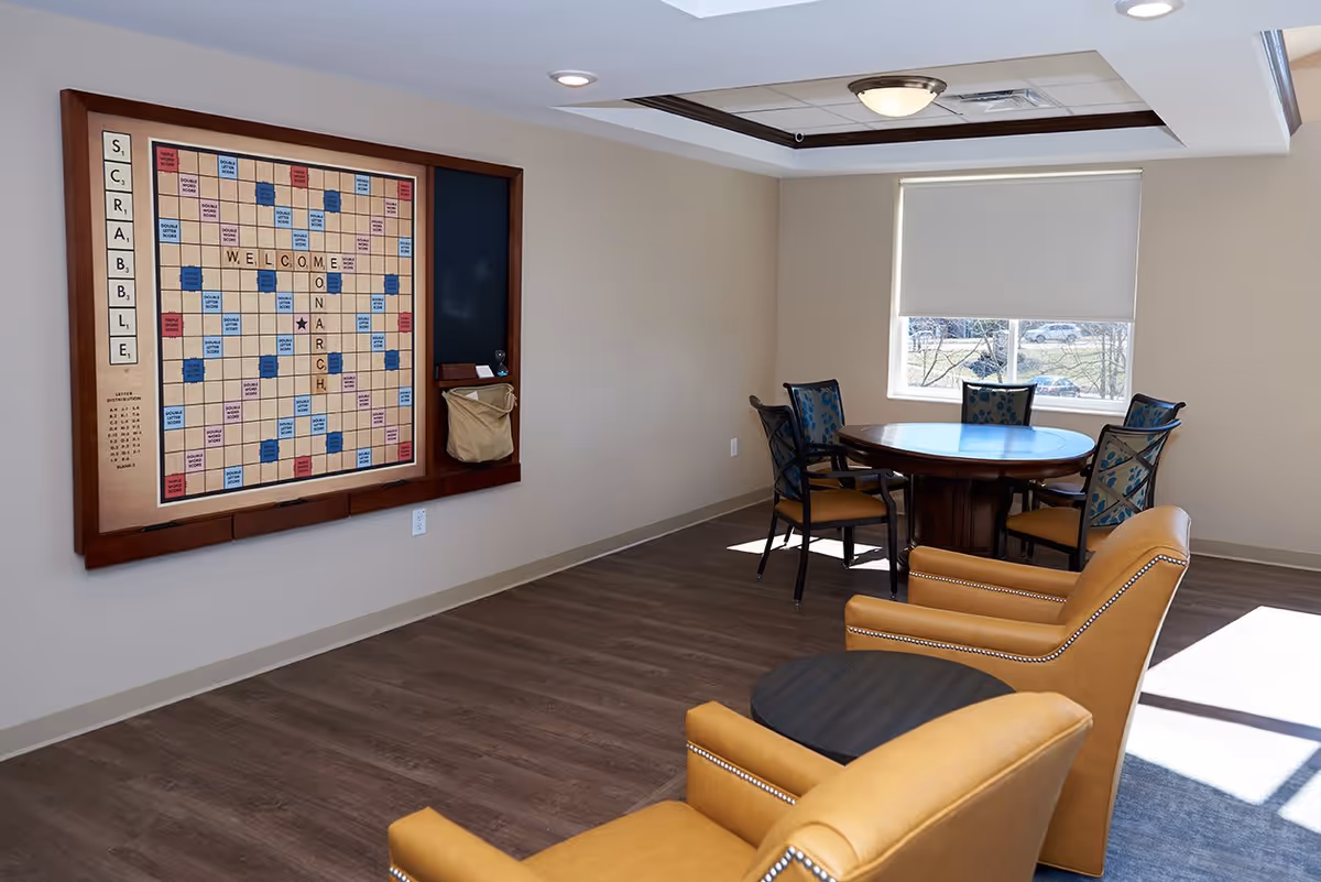 Bright common room with a large wall-mounted Scrabble board, a round table with chairs, and two tan armchairs near a window.