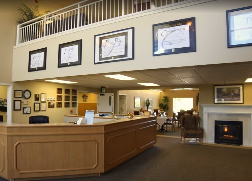 Reception area of Marbella Oroville facility with a wooden front desk, framed certificates on the wall above, a fireplace with a painting above it, and a seating area with chairs and tables in the background.