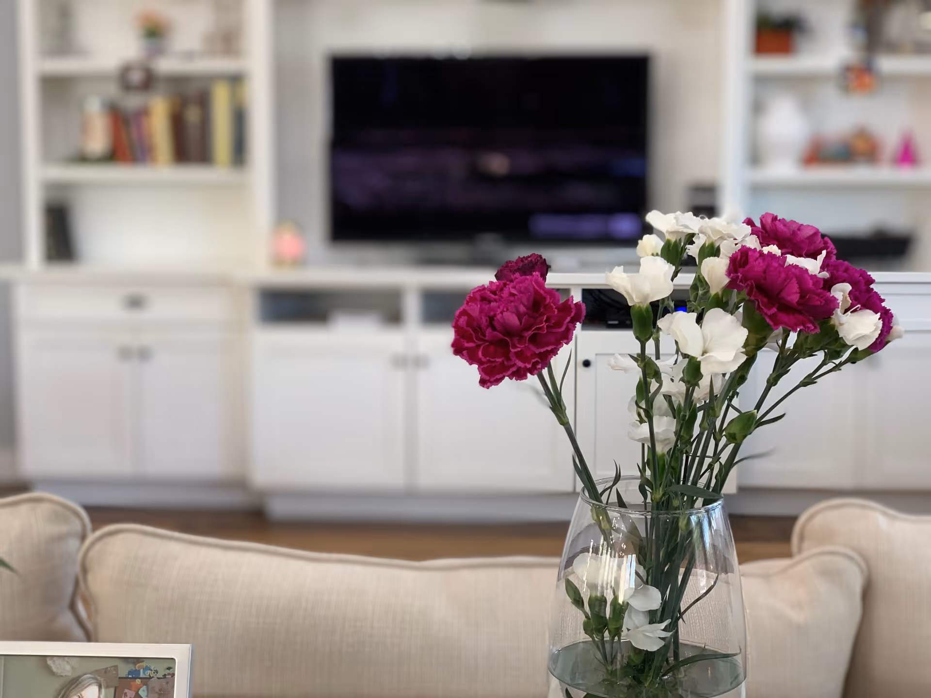 Vase of pink and white flowers on a table in front of a sofa with built-in white cabinets and a TV in the background.