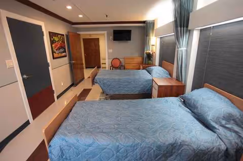 Twin hospital-style beds with blue bedding, curtains, dresser, chair, and wall-mounted TV in a shared room.