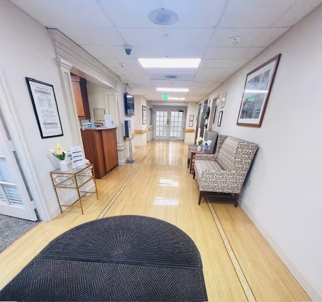 A clean, well-lit hallway in a senior living facility with light wood flooring and white walls. On the right side, there is a patterned bench with a small table holding a flower vase and framed photos. The left side features a small table with a flower vase and informational materials. The hallway leads to double glass doors at the far end, with framed pictures hanging on the walls and ceiling lights illuminating the space.