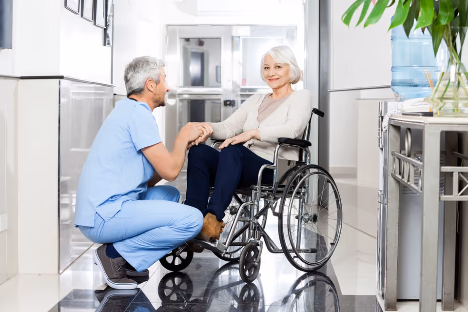 A healthcare worker in blue scrubs kneels and holds hands with an elderly woman sitting in a wheelchair in a bright hallway of a care facility. The woman is smiling and the setting appears clean and welcoming.