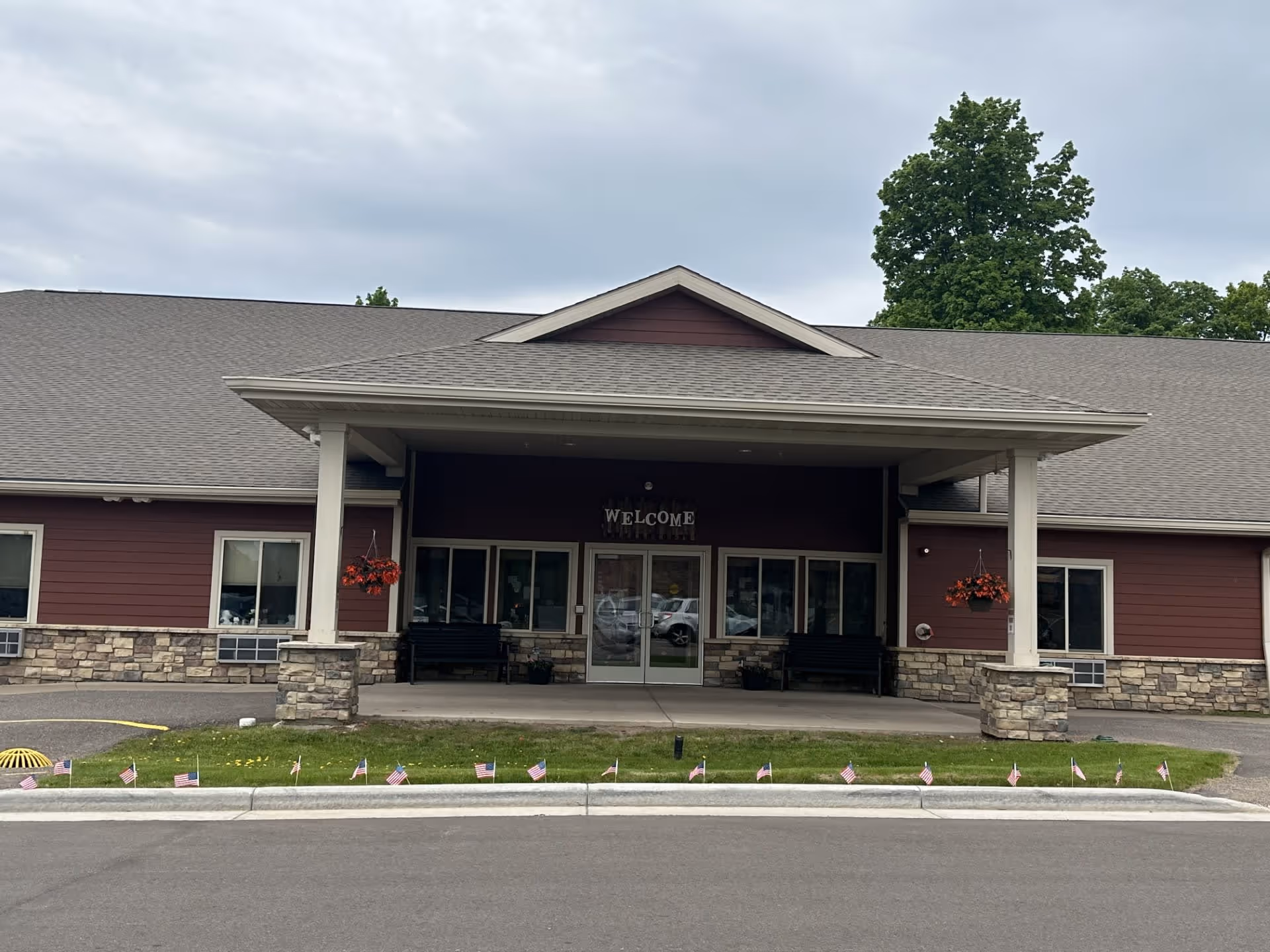 Front exterior view of The Waterford at Park Falls building with a covered entrance, two benches, hanging flower baskets, and small American flags lining the curb in front of the entrance.