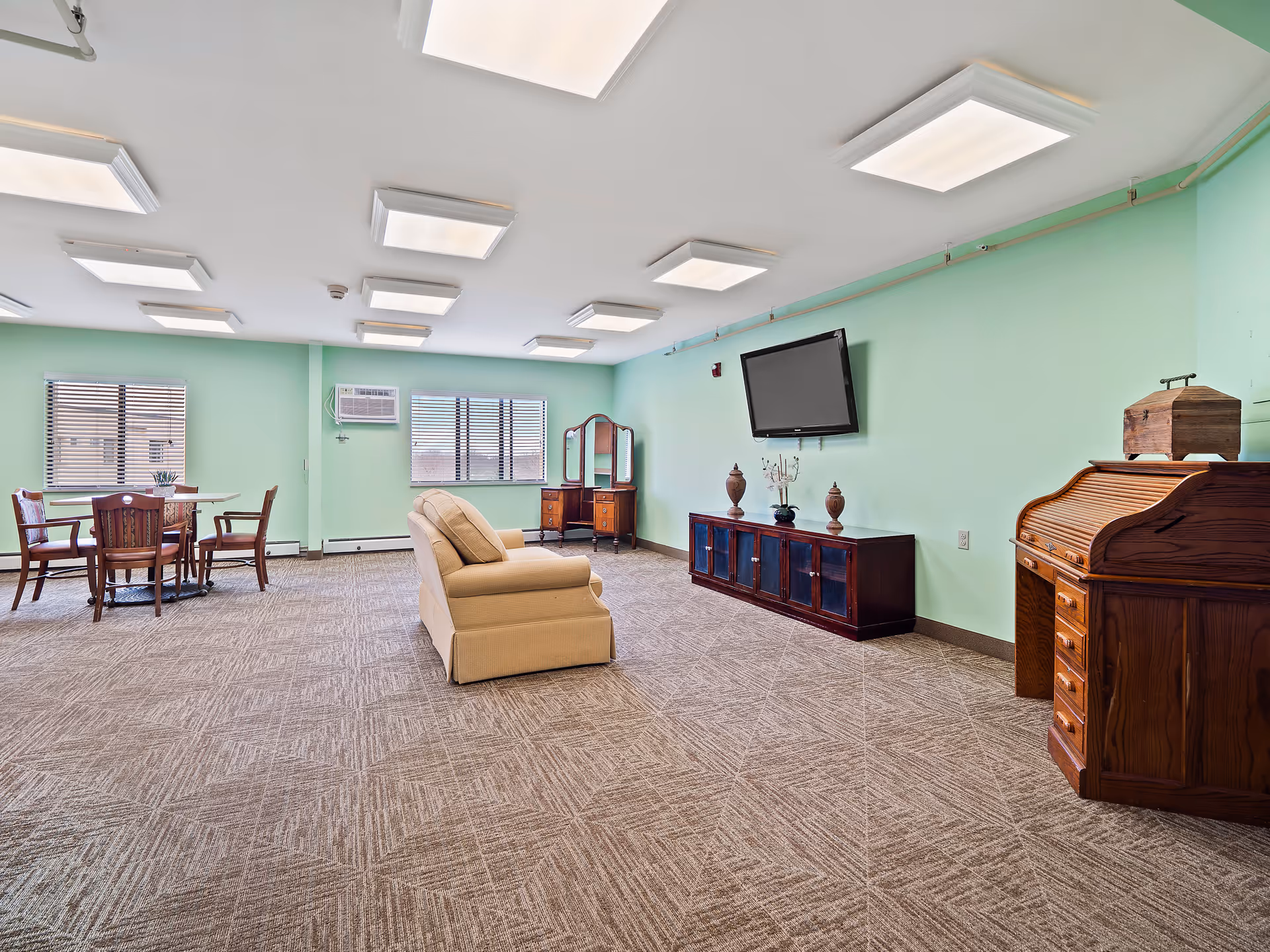 A spacious room with light green walls and patterned carpet flooring. The room features a beige upholstered armchair in the center, a wooden roll-top desk on the right, a dark wooden cabinet with decorative items and a wall-mounted flat-screen TV, a wooden vanity with a mirror near the windows, and a round wooden table with four chairs near two windows with blinds. The ceiling has multiple rectangular light fixtures.