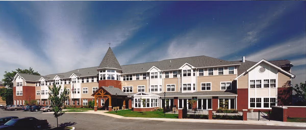 Exterior view of Munhall Retirement Residence, a large multi-story building with a peaked roof and multiple windows, surrounded by a parking lot and some greenery under a partly cloudy sky.