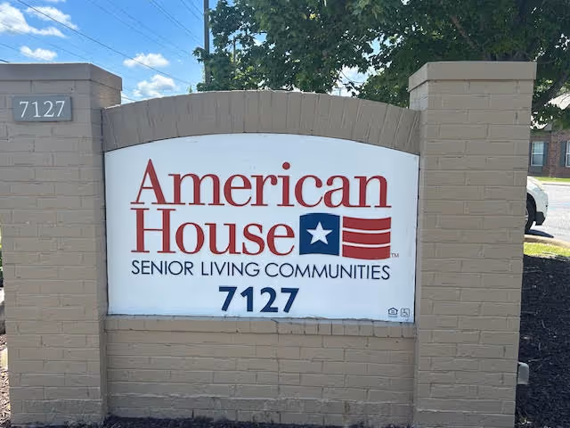 Outdoor sign for American House Senior Living Communities with the address number 7127 displayed on a brick structure, with trees and a street visible in the background.