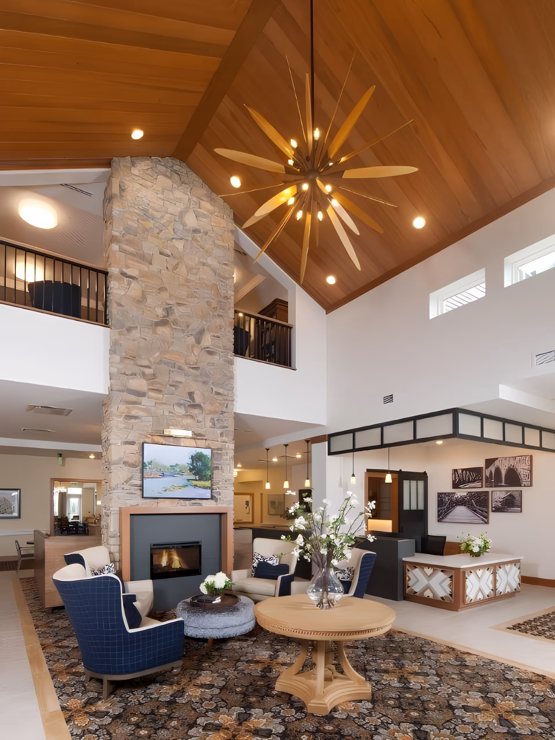 A spacious and elegant living room area with a high wooden ceiling and a large stone fireplace. The room features comfortable seating including blue and cream armchairs arranged around a round wooden table and a small ottoman. A modern chandelier with wooden blades hangs from the ceiling. The fireplace has a TV mounted above it displaying a nature scene. The room is decorated with floral arrangements and has a patterned carpet. In the background, there is a reception desk and framed artwork on the walls.