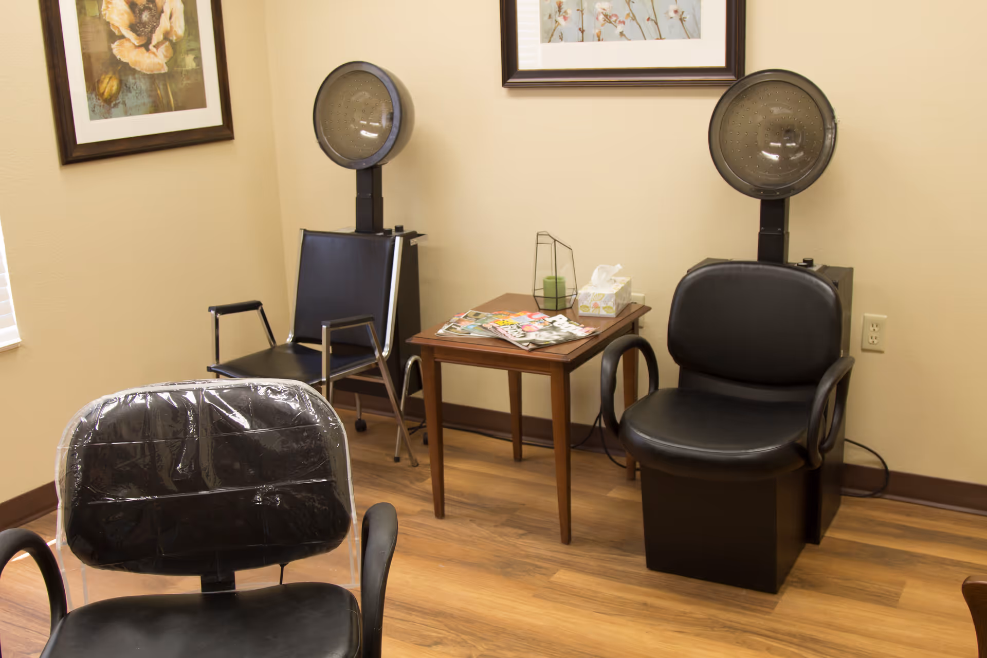 Small salon room with black styling chairs and hooded hair dryers around a side table with magazines.