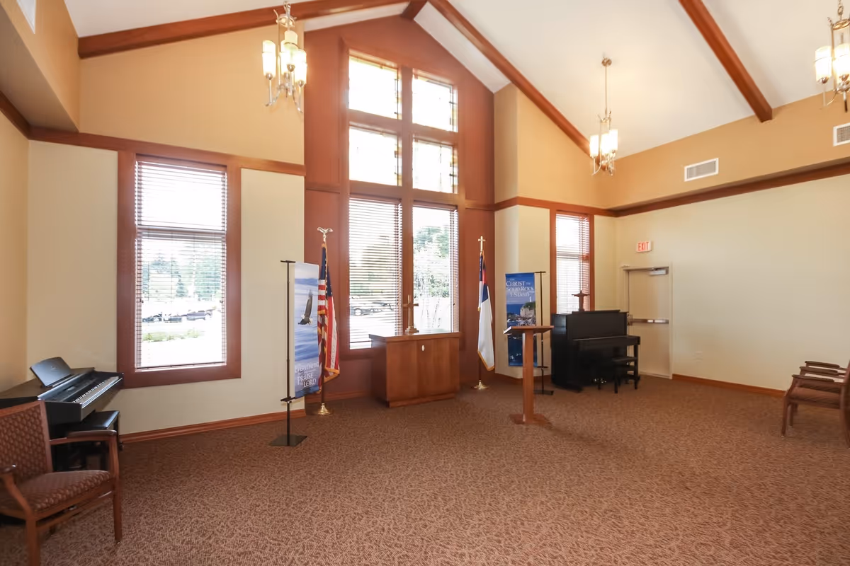 A spacious room with high vaulted ceilings and large windows with wooden frames. The room contains a piano, a keyboard, several chairs, two flags, a wooden podium, and banners with religious themes. The walls are painted beige with wooden trim, and there are multiple hanging light fixtures.