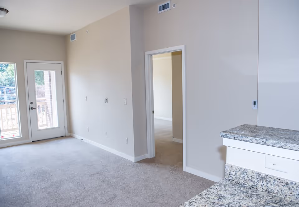 Empty interior room with beige walls and carpeted floor, a door with glass panels leading outside, a window beside the door, and a granite countertop on the right side. There is an open doorway leading to another room.