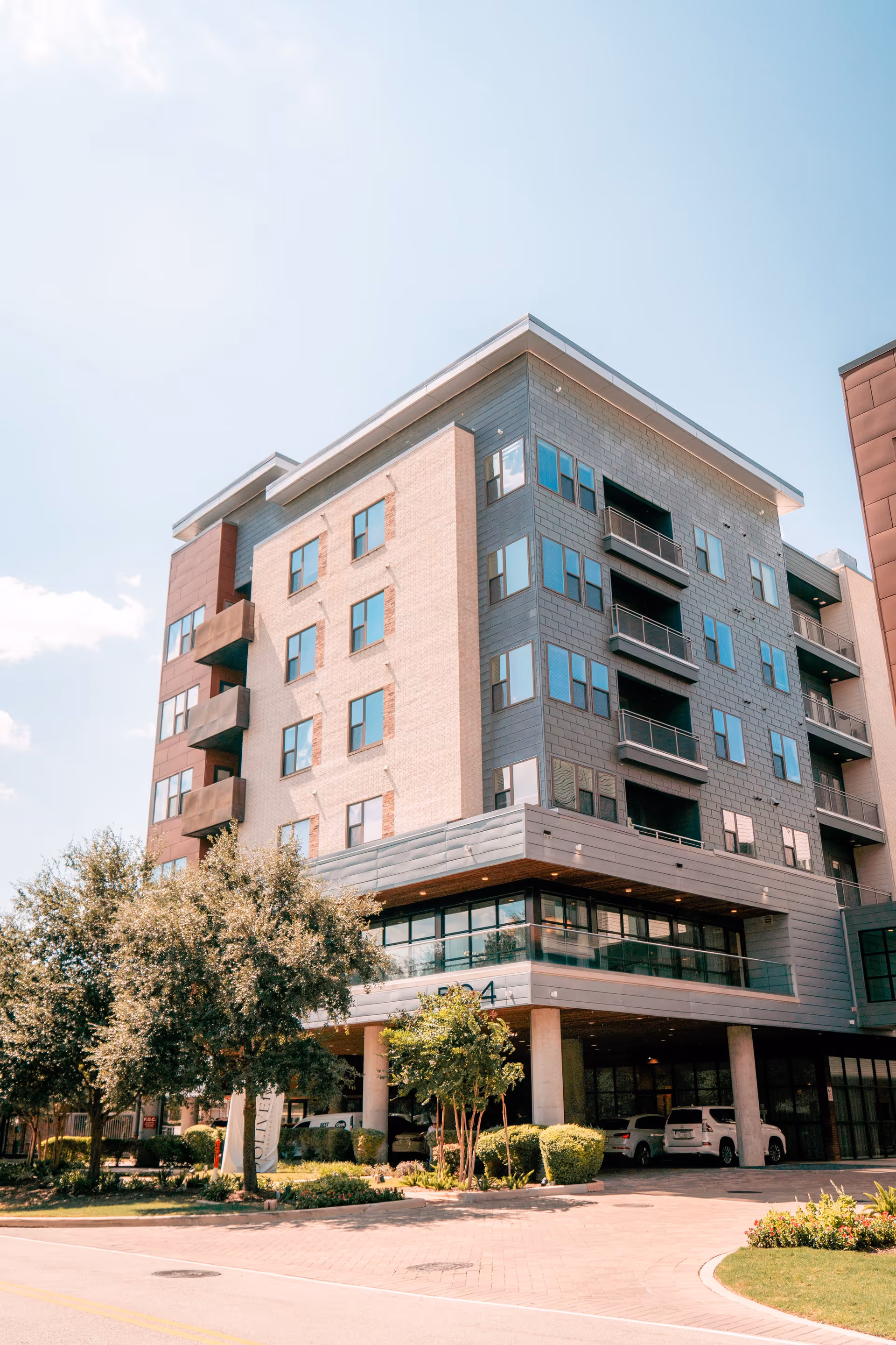 A modern multi-story building with large windows and balconies, surrounded by trees and greenery under a clear blue sky.