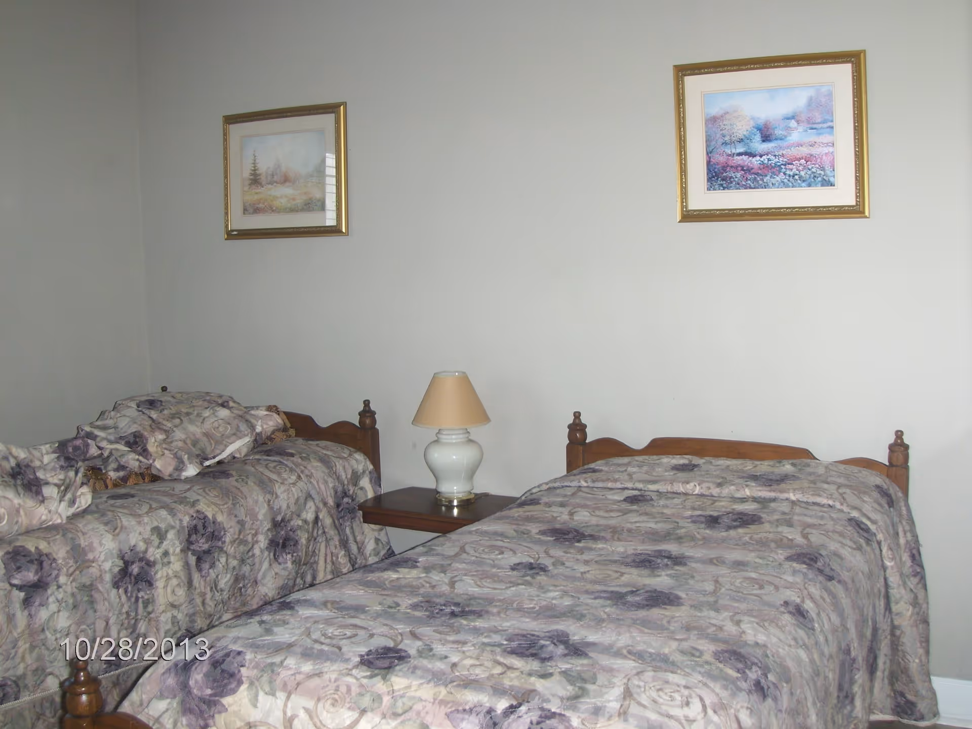 A bedroom with two single beds covered in floral patterned bedspreads in shades of purple and beige. A small wooden nightstand with a white lamp and beige lampshade is placed between the beds. Two framed landscape paintings hang on the light-colored wall above the beds.