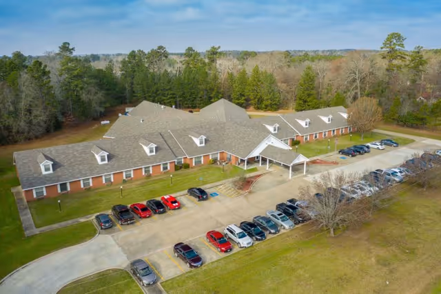 Aerial view of a single-story brick healthcare and rehabilitation center building with a gray shingled roof, surrounded by trees and a parking lot filled with cars. The building has multiple dormer windows and a covered entrance.