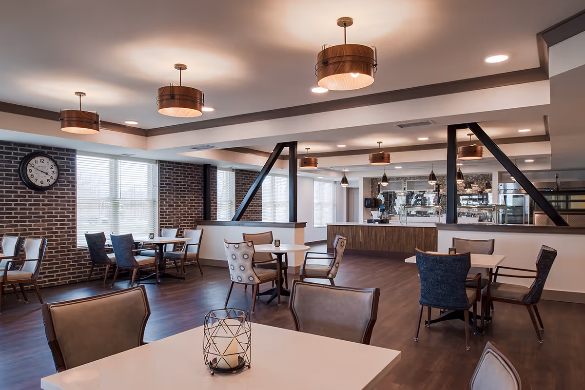 A spacious dining room with multiple tables and chairs arranged neatly. The room features large windows with blinds, exposed brick walls, modern pendant lighting, and a clock on the wall. The far end has a counter area with kitchen equipment visible behind it.