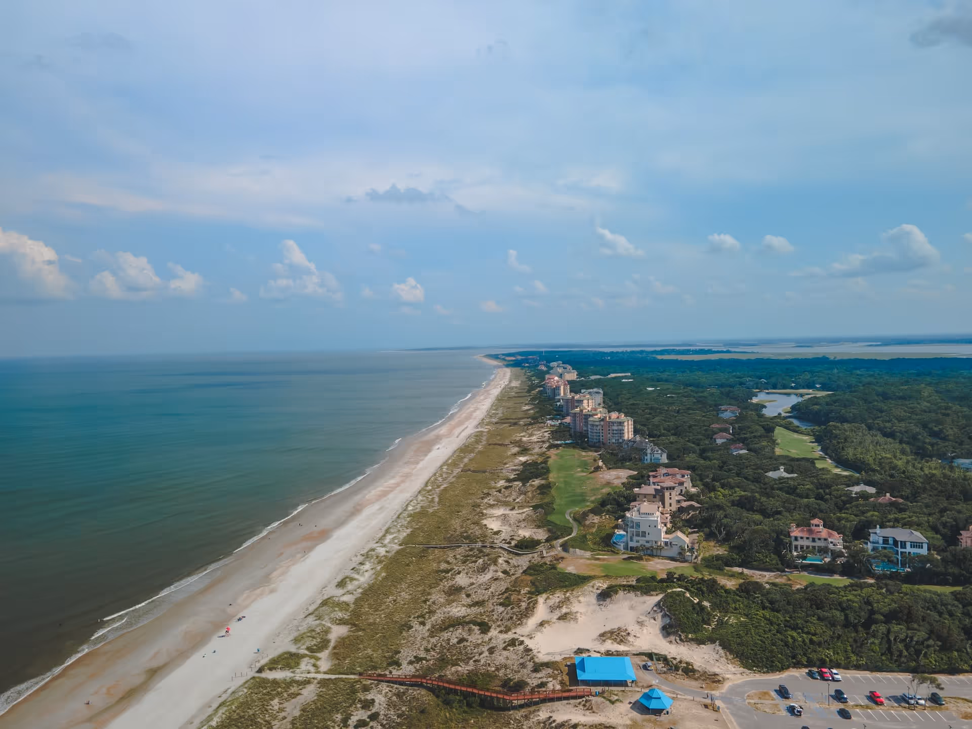 Aerial view of a long sandy beach stretching along the coastline with calm ocean waters on the left and a row of residential buildings and greenery on the right under a partly cloudy sky.