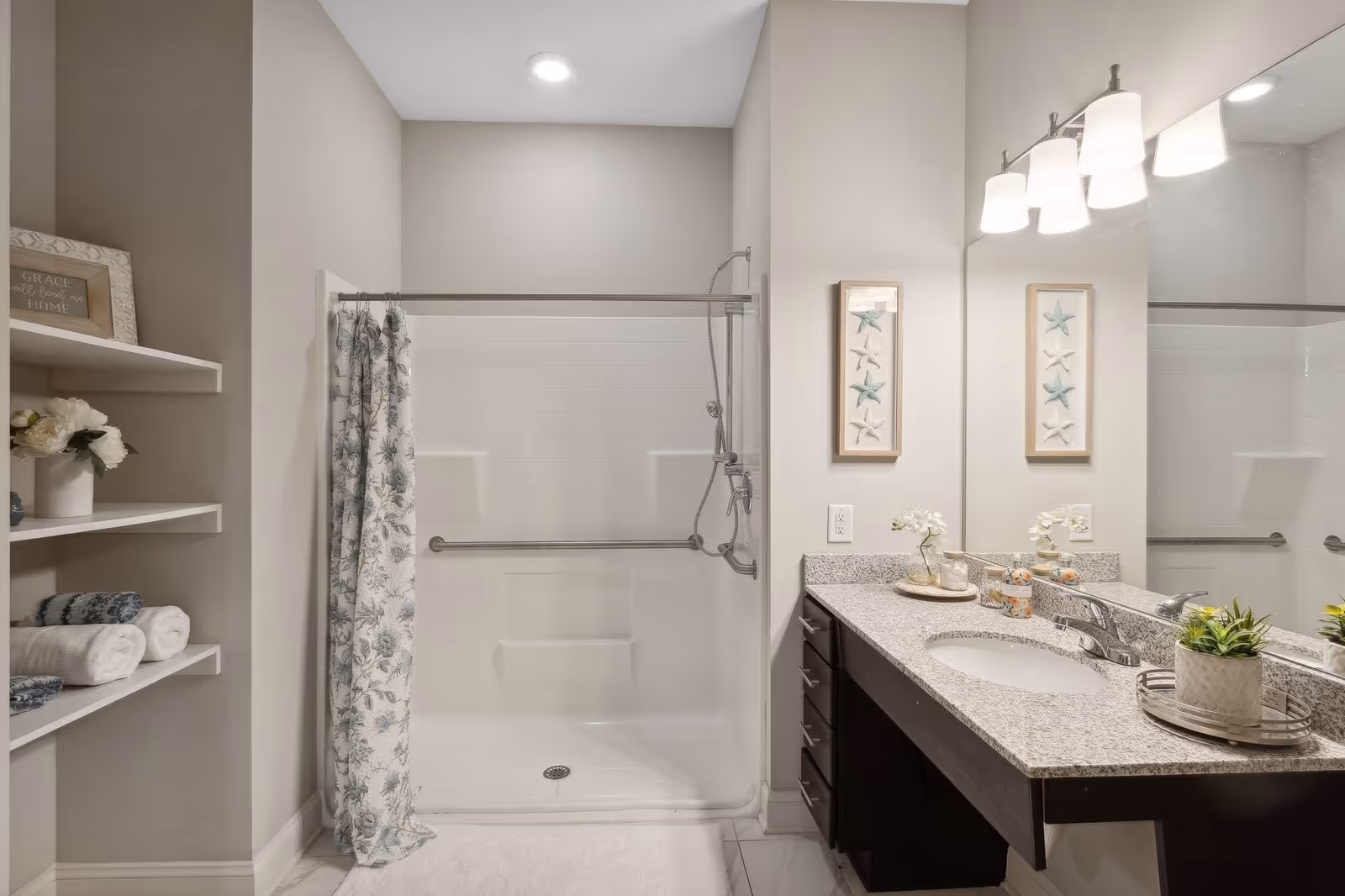 A modern bathroom featuring a walk-in shower with a floral shower curtain, a granite countertop with a sink, a large mirror, and decorative items including framed starfish artwork, a small plant, and neatly folded towels on white shelves.