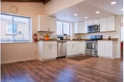 A modern kitchen with white cabinets, stainless steel appliances including a microwave and oven, a dishwasher, and a sink. The kitchen has wooden flooring and two windows letting in natural light. A fire extinguisher is mounted on the wall near one window.