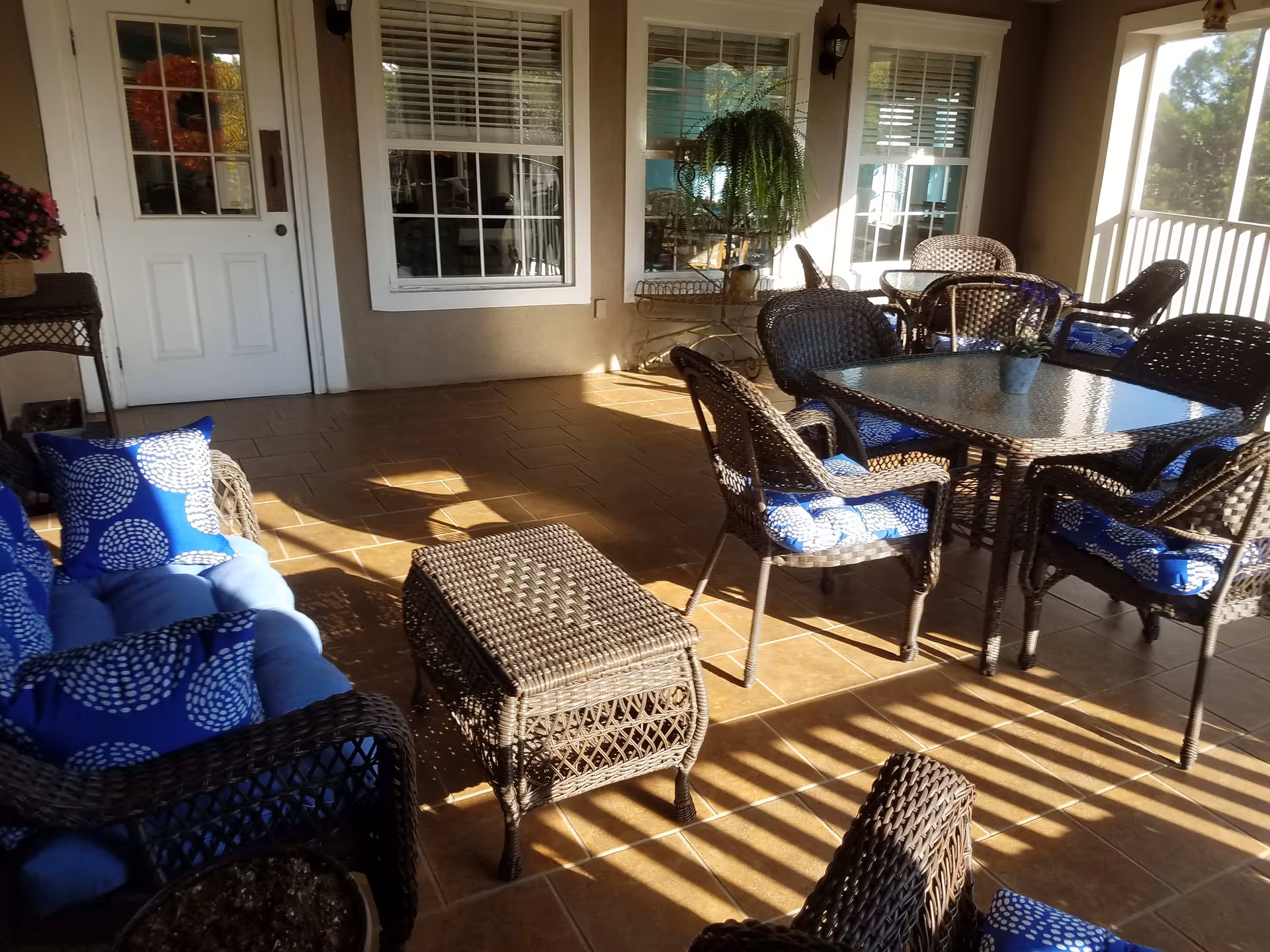 A sunlit outdoor patio area with wicker furniture including a sofa, chairs with blue patterned cushions, a glass-top table, and a small wicker coffee table. The patio has tiled flooring and is adjacent to a building with windows and a door. There is a hanging fern plant and a small potted plant on the table.