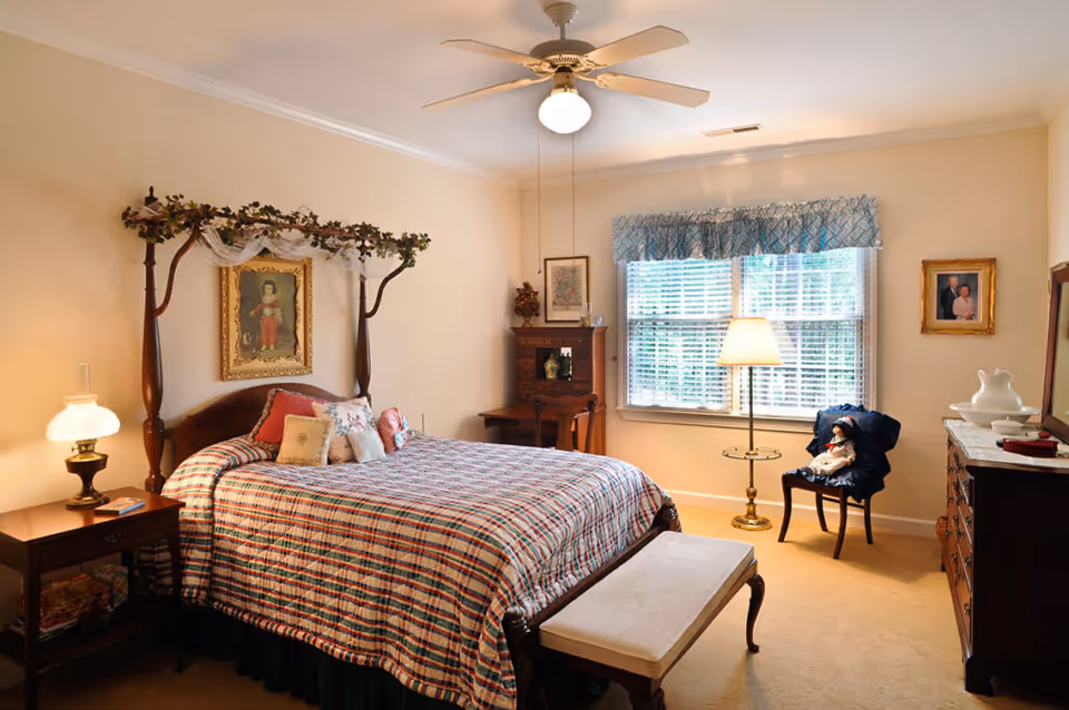 A cozy bedroom with a wooden four-poster bed adorned with a plaid bedspread and decorative pillows. There is a wooden nightstand with a lamp and a book, a bench at the foot of the bed, a wooden dresser with a mirror, and a chair with a doll sitting on it. The room has beige walls, a ceiling fan with a light, and a window with blue valance curtains letting in natural light.