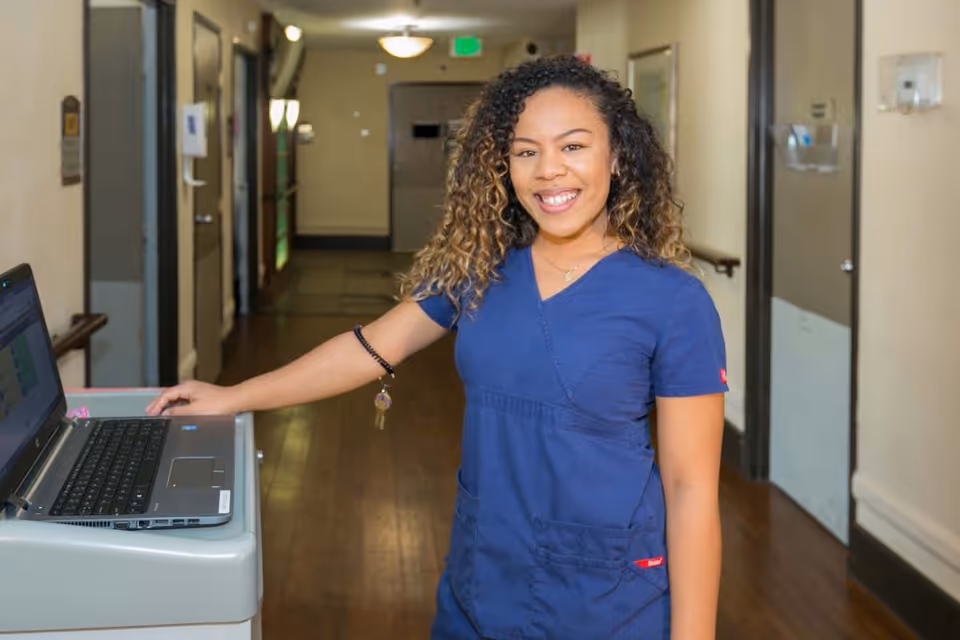 A smiling healthcare worker in blue scrubs standing in a hallway next to a mobile computer workstation with a laptop on it.