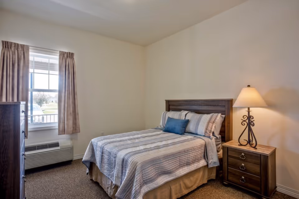 A bedroom with a neatly made bed featuring striped bedding and a blue accent pillow. Next to the bed is a wooden nightstand with a decorative lamp. There is a window with beige curtains and a wooden dresser near the window. The walls are plain and light-colored, and the floor is carpeted.