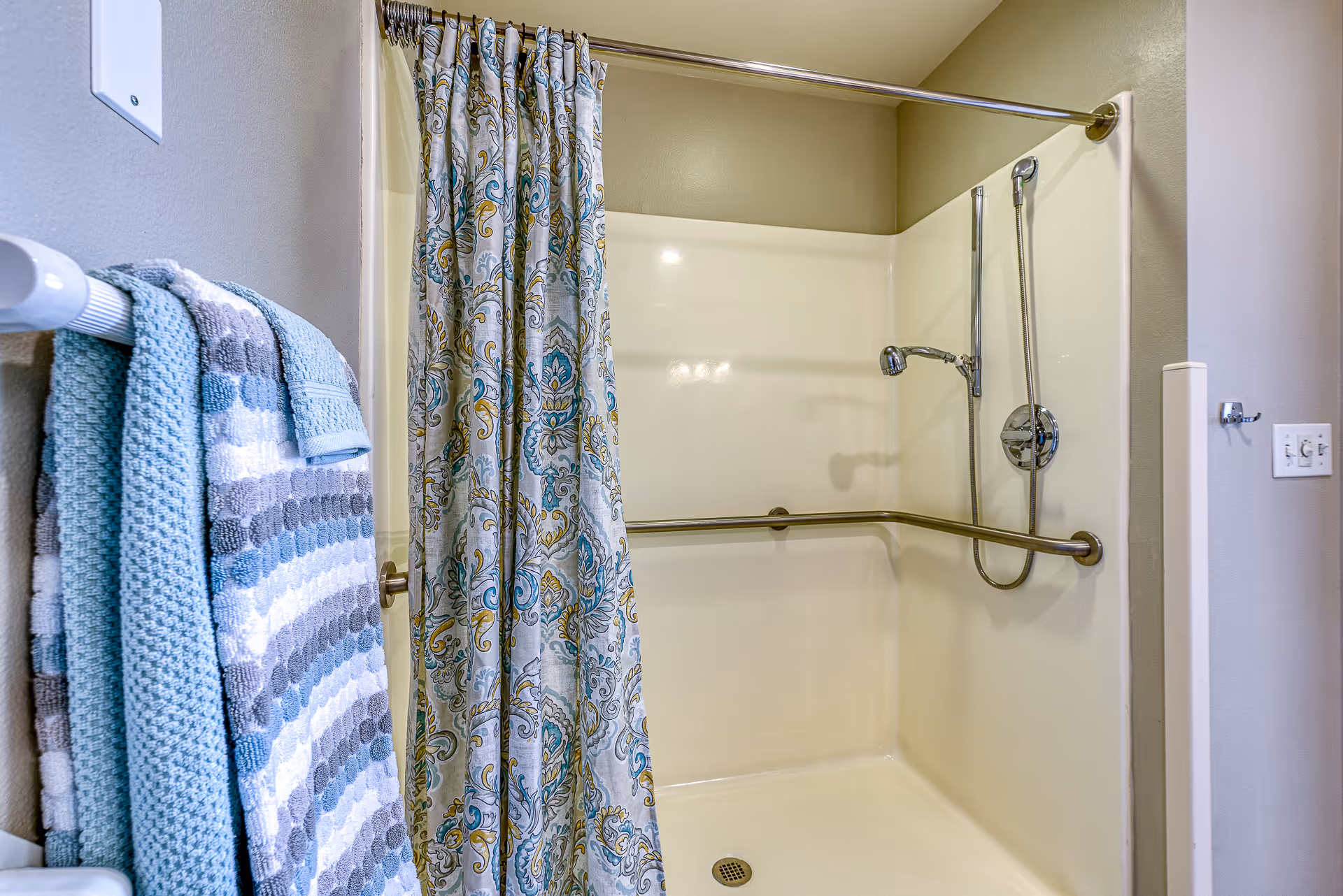 A bathroom shower area with a white shower base and walls, a silver showerhead with a handheld attachment, and a horizontal grab bar. A patterned shower curtain with blue, yellow, and gray designs hangs on a silver rod. On the left side, several folded towels in shades of blue, gray, and white are hanging on a towel rack.