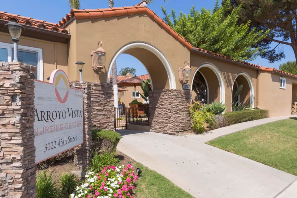Exterior view of Arroyo Vista Nursing Center showing a beige building with red tile roof, stone pillars, arched entryways, a gated entrance, and a sign with the facility name and address. There are green plants, flowers, and a clear blue sky.