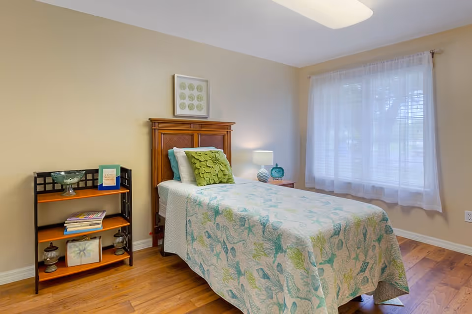 A cozy bedroom with a single bed featuring a wooden headboard, a green decorative pillow, and a light blue and green patterned quilt. Next to the bed is a wooden nightstand with a lamp and a blue clock. A window with sheer white curtains allows natural light to fill the room. There is a small black and wooden shelf with books and decorative items against the beige wall. The floor is wooden.