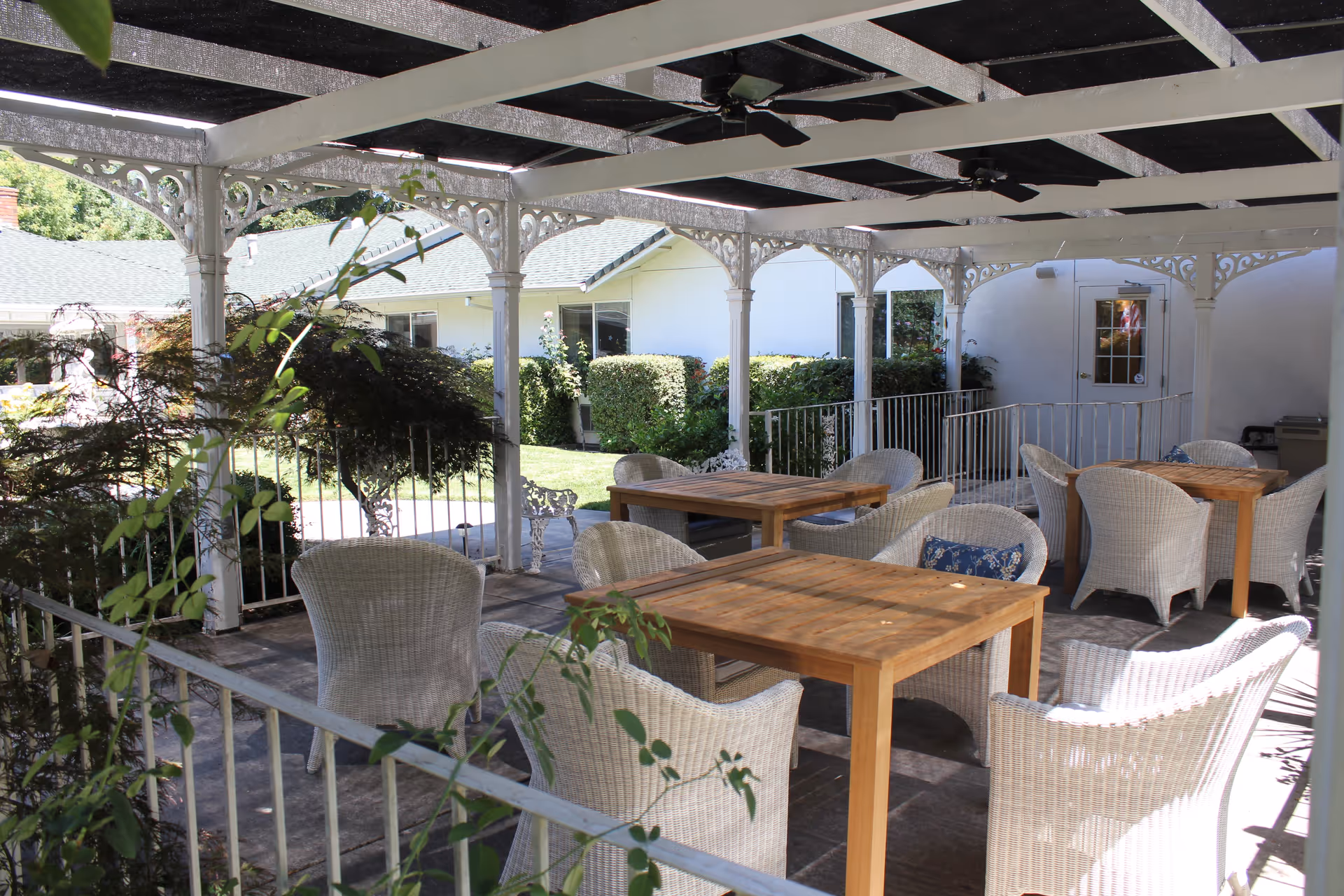 Outdoor covered patio area with wooden tables and white wicker chairs arranged for seating. The patio has decorative white columns and a ceiling fan, with greenery and bushes visible in the background along with a white building.