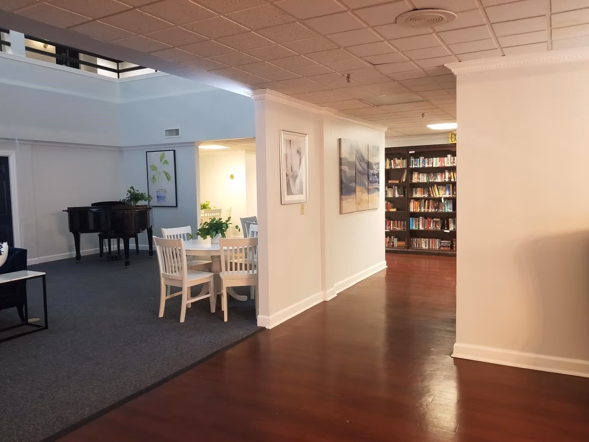 Interior view of a senior living facility showing a hallway with wooden flooring leading to a room with bookshelves filled with books. To the left, there is a carpeted area with a round table surrounded by white chairs and a black grand piano in the background. The walls are decorated with framed artwork and there are plants on the table and near the piano.
