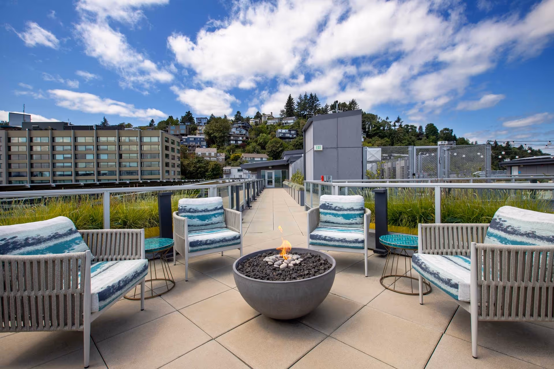 Outdoor patio area with four cushioned chairs arranged around a circular fire pit. The patio has tiled flooring and glass railings with greenery behind them. In the background, there are buildings and a hillside with houses under a partly cloudy blue sky.