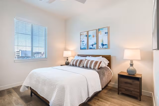 A bright bedroom with a large bed covered in white and brown bedding, two bedside tables each with a lamp, three abstract paintings on the wall above the bed, and a window with blinds letting in natural light.