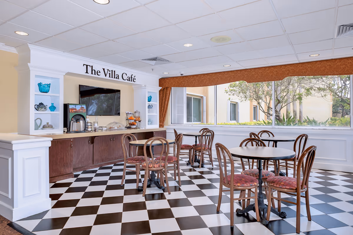 Interior view of The Villa Café with black and white checkered floor, round tables with wooden chairs that have red cushioned seats, a counter with a coffee machine, fruit dispenser, and decorative shelves with vases. Large windows show trees and a building outside.