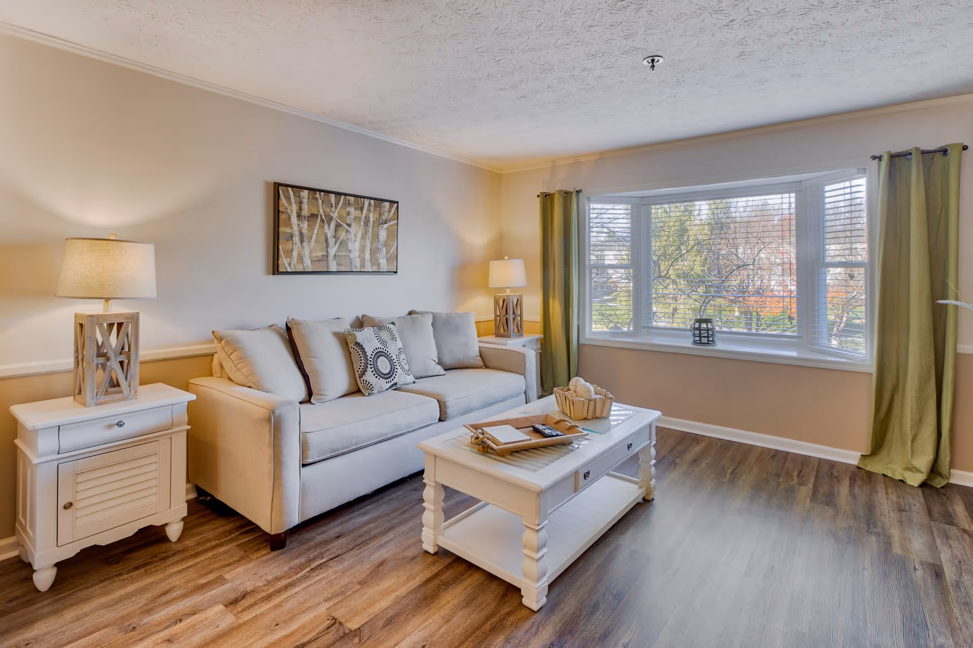 A cozy living room with a beige sofa adorned with cushions, two white side tables each with a lamp, a white coffee table with decorative items, and a large bay window with green curtains letting in natural light. The room has wooden flooring and a framed artwork of birch trees on the wall.