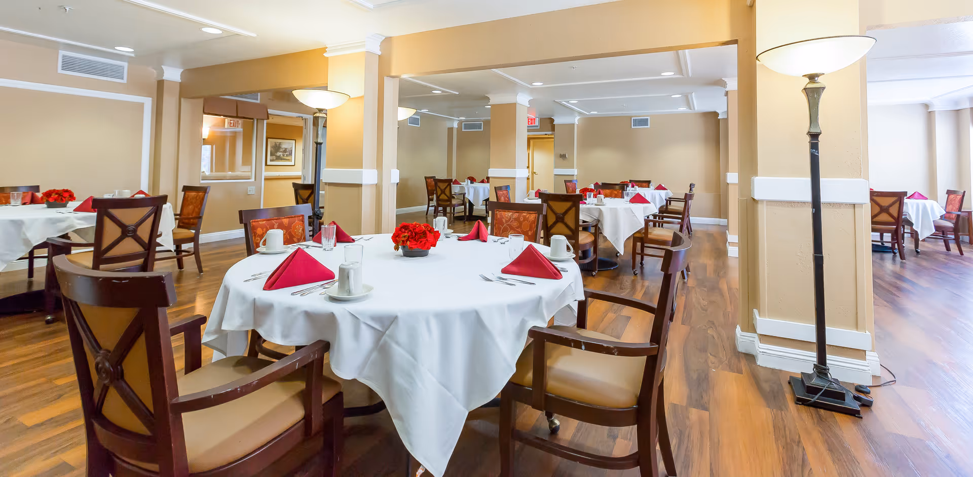 Elegantly set dining room with round tables draped in white linens, red napkins, floral centerpieces, and wooden chairs on hardwood floors.