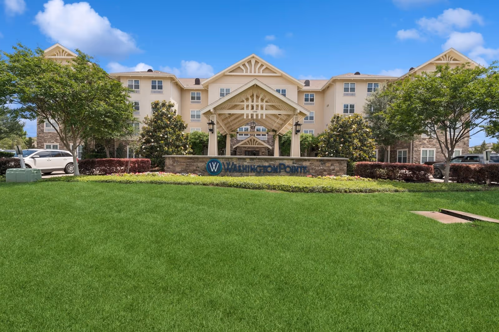 Front exterior view of the Washington Pointe senior living facility with a well-maintained green lawn, trees, and a covered entrance structure under a blue sky with some clouds.