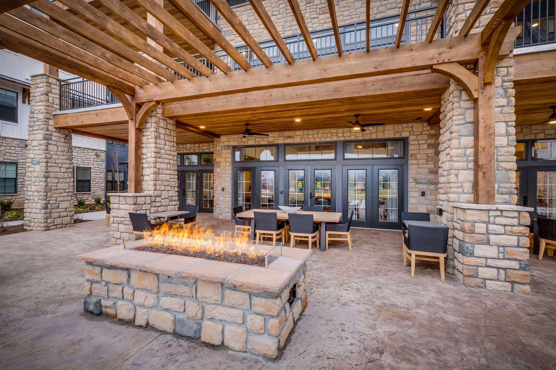 Outdoor patio area at Everlan of Hixson featuring a stone fire pit with flames, wooden pergola overhead, stone pillars, and several tables and chairs for seating. The background shows large glass doors leading inside the building.