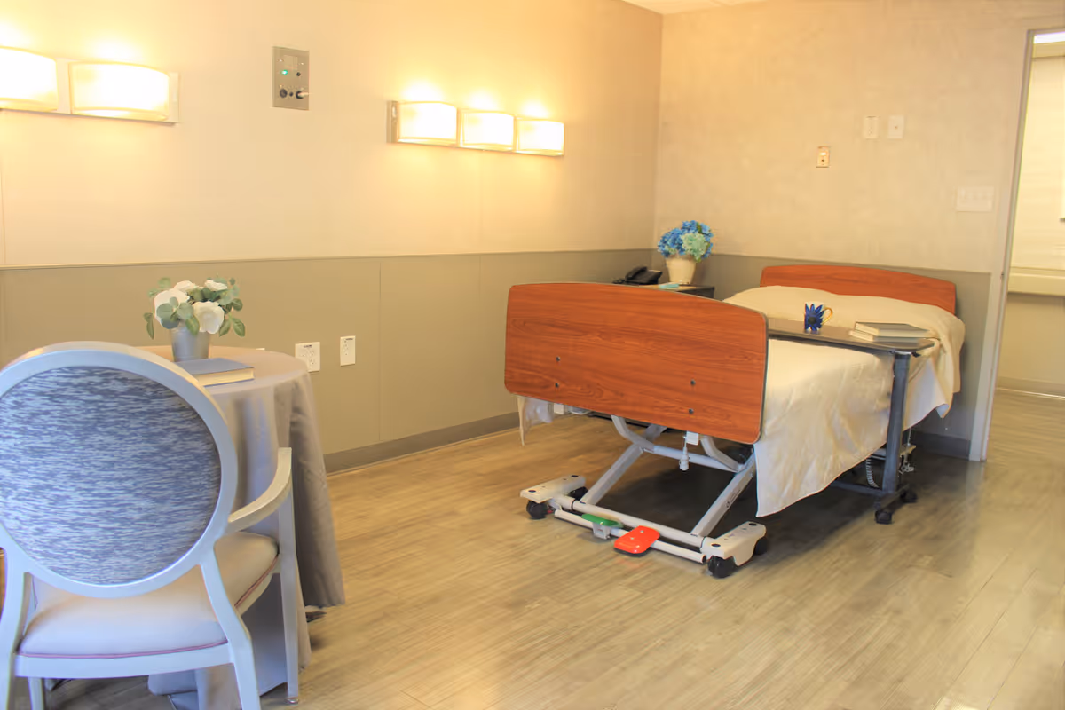A well-lit room with a hospital-style adjustable bed covered with a beige blanket, a small bedside table with a blue flower vase and books, and a round table with a gray tablecloth and a chair. The walls have mounted lights and the floor is wooden.