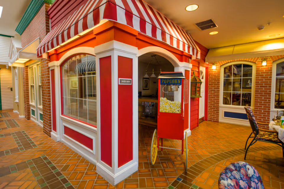 Interior view of a senior living facility featuring a colorful corner shop facade labeled 'Sweet Shop' with a red and white striped awning. In front of the shop is a vintage-style popcorn machine filled with popcorn. The floor is tiled with a brick pattern, and there are chairs with floral upholstery and tables nearby. The walls have a mix of brick and painted surfaces with windows and warm lighting.