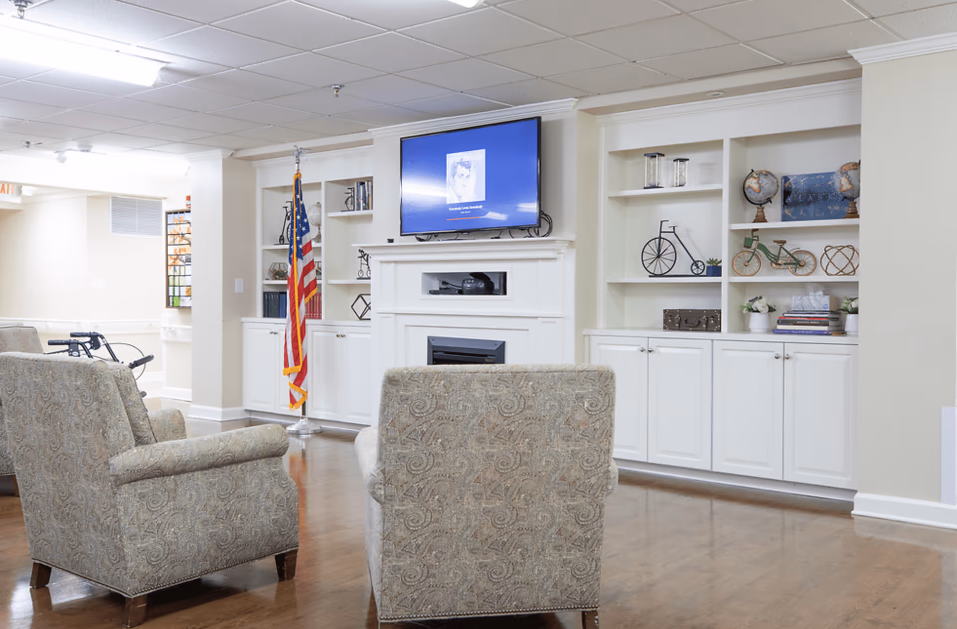 A cozy living room area in a senior living facility with two patterned armchairs facing a wall-mounted TV above a white fireplace. The wall behind the fireplace has built-in white shelves decorated with globes, books, and decorative items. An American flag stands to the left of the fireplace. The room has wooden flooring and a drop ceiling with fluorescent lighting.