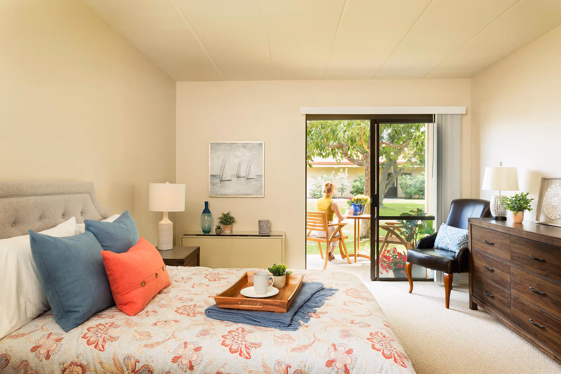 A bright and cozy bedroom with a bed featuring floral bedding and colorful pillows. There is a wooden dresser with a lamp and plant on top, a black armchair with a decorative pillow, and a sliding glass door leading to an outdoor patio where a person is sitting at a table surrounded by greenery.