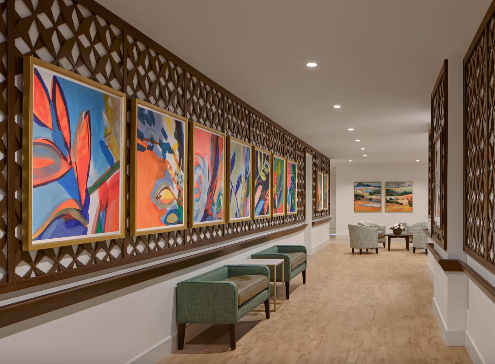A well-lit hallway in The 501 at Mattison Estate featuring a series of colorful abstract paintings mounted on a decorative wooden lattice wall. The hallway has light wood flooring, two green upholstered benches with small tables between them along one side, and a seating area with four patterned armchairs and a round wooden table with a flower arrangement at the far end.