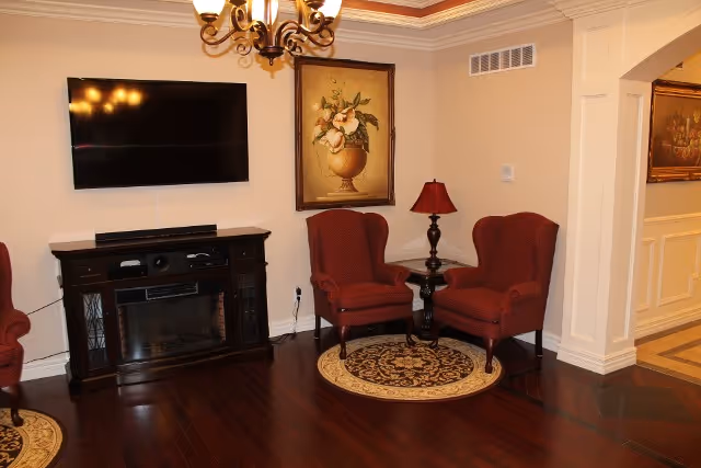 A cozy living room area with two red upholstered armchairs placed on a round patterned rug. Between the chairs is a small dark wooden side table with a red lampshade. On the wall above the chairs is a framed painting of flowers in a vase. To the left, a flat-screen TV is mounted on the wall above a dark wooden media console with a built-in electric fireplace. The room has hardwood flooring and cream-colored walls with crown molding and recessed lighting.