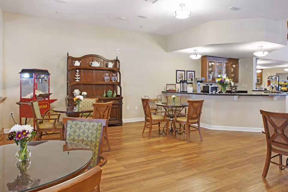 A cozy common area with wooden floors featuring several round tables with chairs, each table decorated with a small vase of flowers. There is a wooden hutch against the wall displaying teapots and glassware. A popcorn machine is positioned near the hutch. In the background, there is a counter area with coffee dispensers, a coffee machine, and a flower arrangement. The space is well-lit with ceiling lights and has a warm, inviting atmosphere.