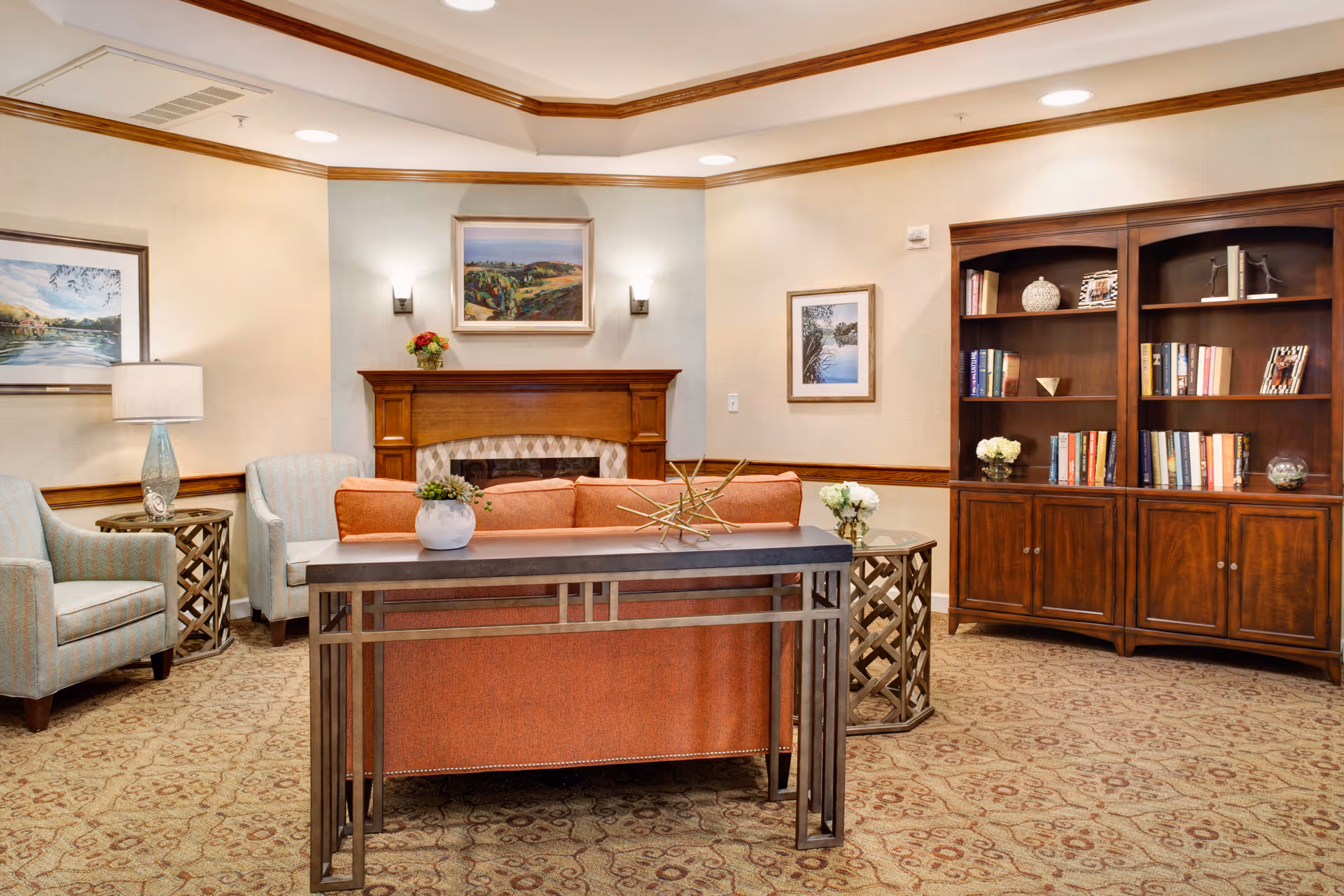 A cozy living room area with a wooden fireplace mantle, an orange sofa, two light blue armchairs, a wooden bookshelf filled with books and decorative items, and framed landscape paintings on the walls. The room has warm lighting and a patterned carpet.