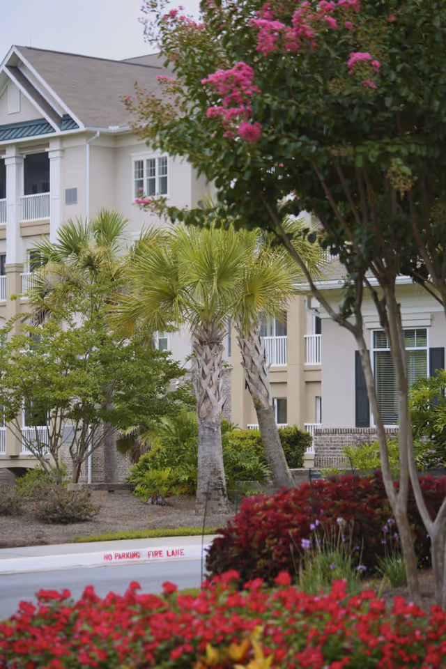 Exterior view of a senior living facility building with balconies, surrounded by palm trees, flowering bushes, and other greenery. The curb is marked with 'No Parking Fire Lane' in red.