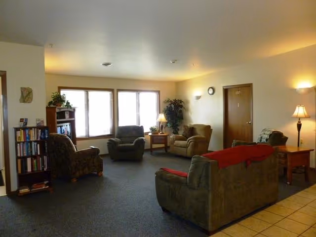Living room in a senior facility with couches, armchairs, lamps, a bookshelf, and large windows.